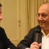 Two men sitting at a table, engaged in conversation, speaking into microphones.